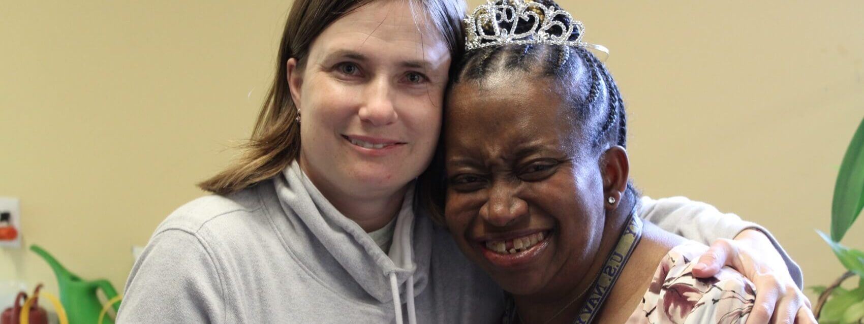 Two women pose together, smiling. One wears a light gray hoodie, the other a tiara and a floral top. They have their arms around each other in a warm, friendly embrace.