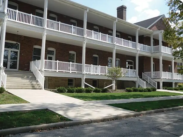 Two-story brick building with white railings, covered porches, exterior staircases on both sides, and a sidewalk in front, surrounded by trimmed bushes and grass.