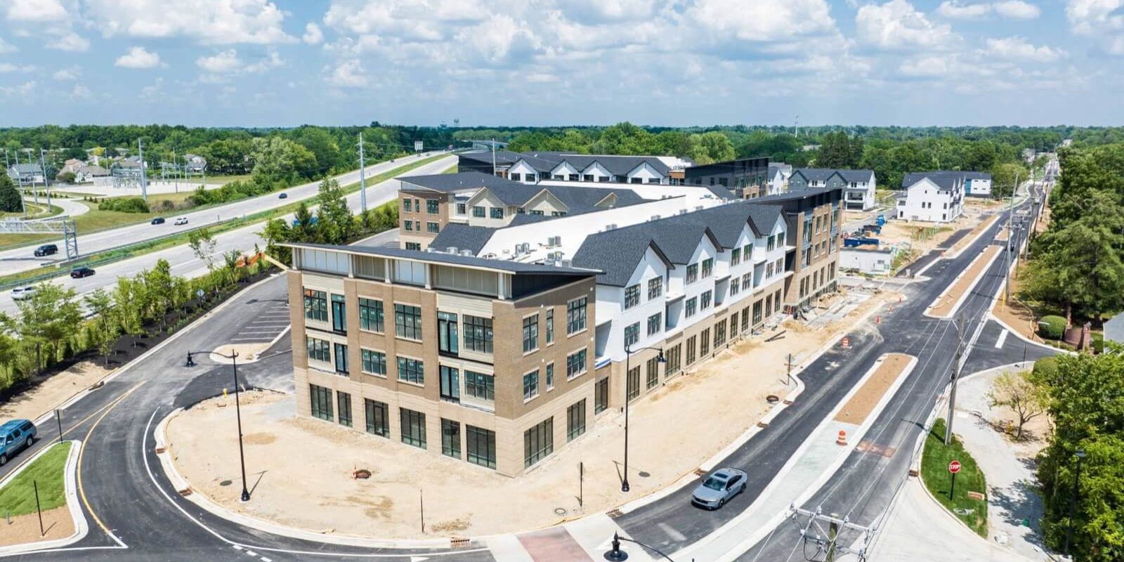 Aerial view of a newly constructed multi-story apartment building, surrounded by freshly paved roads and ongoing landscaping work, with cars and construction cones visible.
