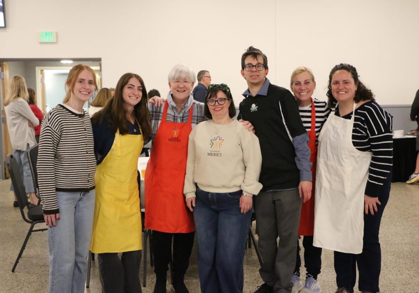 Seven people stand indoors, some wearing aprons, posing and smiling for a group photo in a well-lit room with tables and other people in the background.