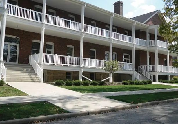Two-story brick building with white railings, covered porches, exterior staircases on both sides, and a sidewalk in front, surrounded by trimmed bushes and grass.