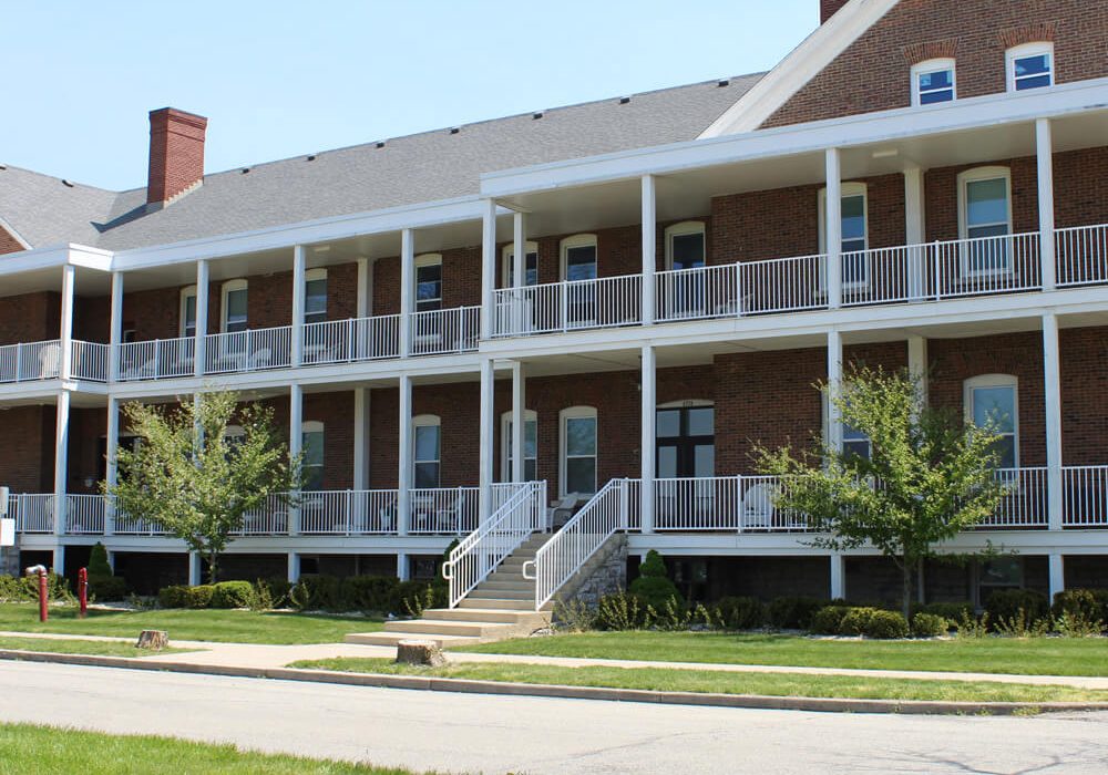 A two-story brick building with white railings and columns, featuring exterior staircases and balconies, is surrounded by grass and small trees.