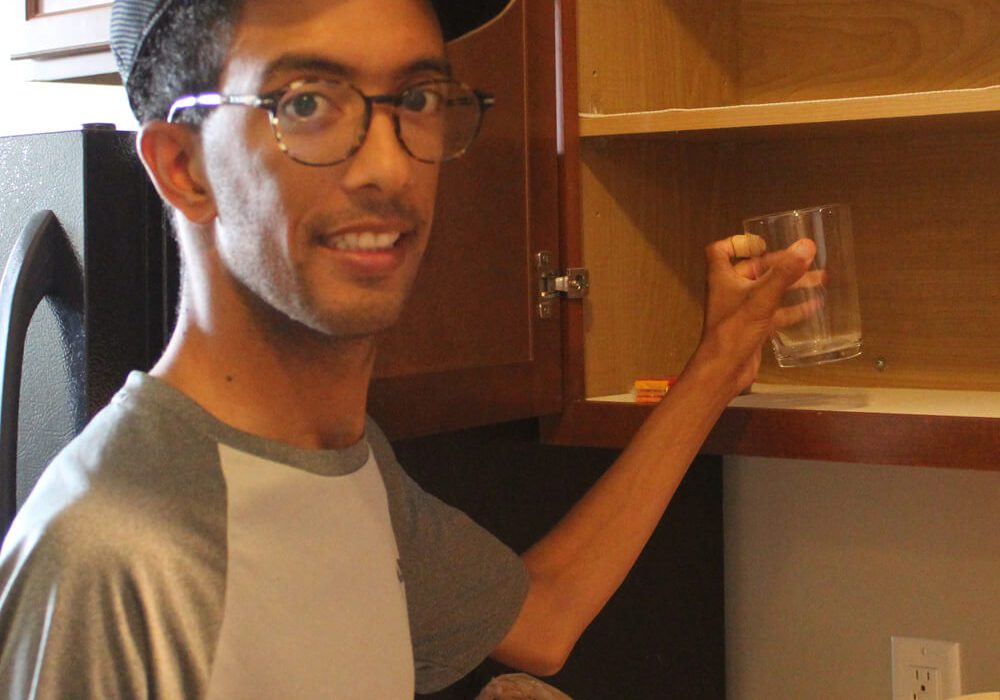 A man wearing glasses and a baseball cap places a clear glass on an empty wooden shelf in a kitchen cabinet.